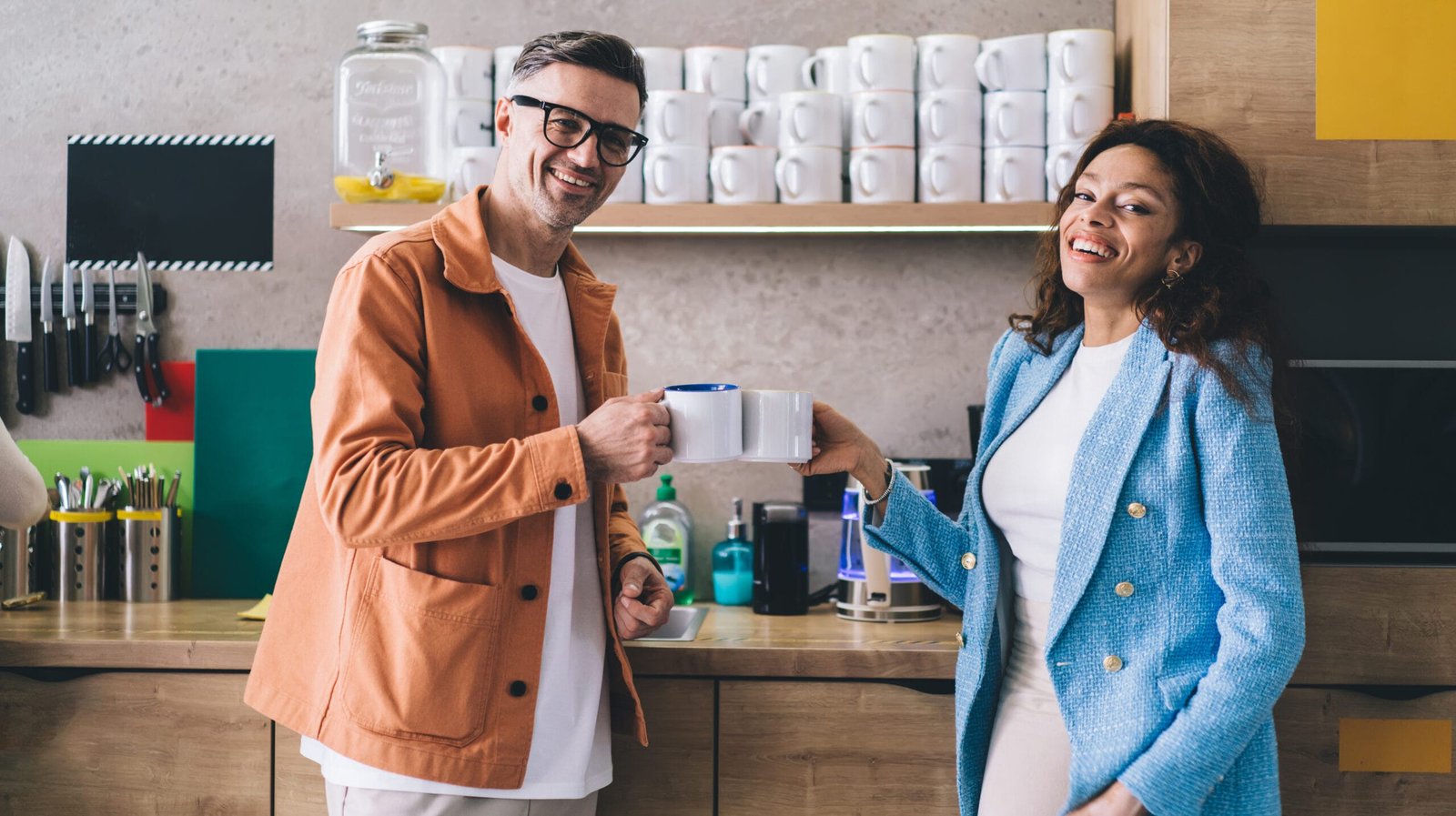 Optimistic adult male and smiling female colleagues in stylish clothes standing near counter in kitchen with cups of coffee while looking at camera