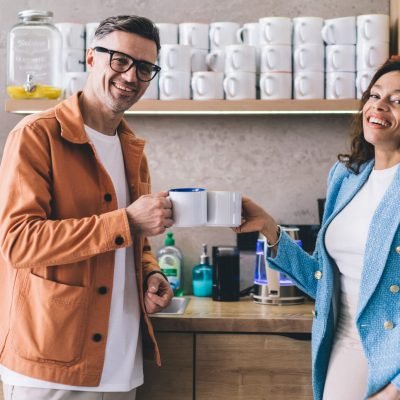 Optimistic adult male and smiling female colleagues in stylish clothes standing near counter in kitchen with cups of coffee while looking at camera